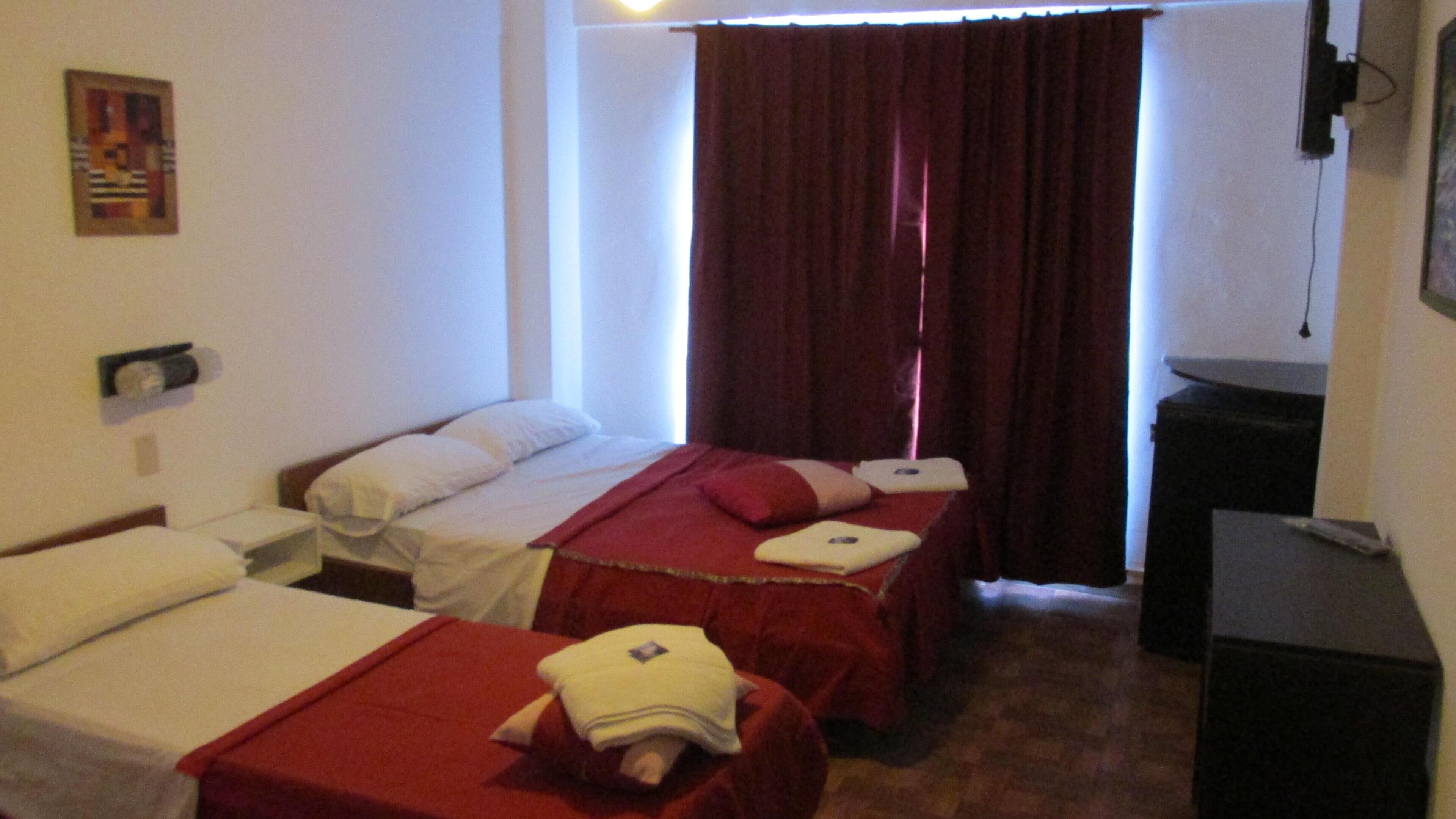 Habitación de hotel con dos camas individuales, un escritorio, una silla y un ventanal con cortinas rojas.