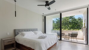 Une chambre moderne avec un grand lit, une table de chevet, un ventilateur de plafond et une porte-fen être coulissante donnant sur un balcon aménagé avec du mobilier d’extérieur.