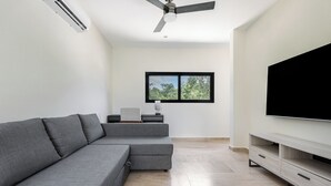 Un salon moderne avec un canapé d’angle gris, un téléviseur à écran plat posé sur un meuble blanc, un ventilateur de plafond et une fenêtre donnant sur la forêt.