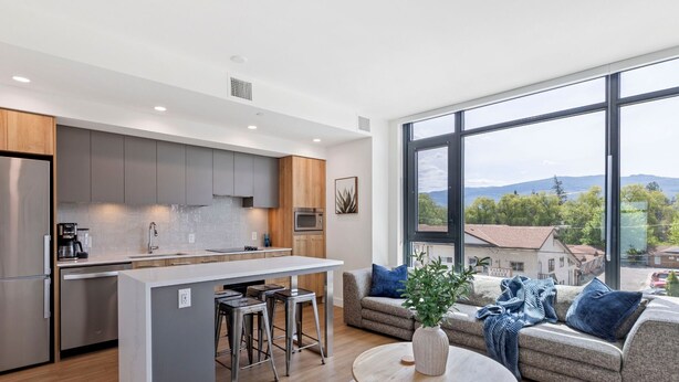 A modern kitchen with a central island, bar stools, and a large window offering a view of a residential area. - Lakeside Retreat w/ Rooftop Pool, Hot Tub, Firepits & BBQs – 10 min to Downtown (Kelowna)