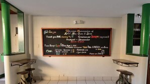 A hotel reception area with a chalkboard displaying breakfast menu and prices, two small tables, and green pillars. - Hotel San Juan de León (León)