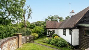 Une maison de campagne traditionnelle, aux murs blancs et au toit en pente, entourée d’un jardin bien entretenu et d’un mur de pierres.