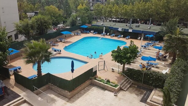 Une piscine dotée de chaises longues et d’une petite cascade, entourée de verdure et avec un bâtiment en arrière-plan.
