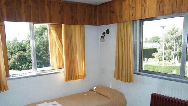 A single bed with a beige bedspread, two windows with yellow curtains, a wall-mounted light fixture, and a radiator. - Residencia Familiar La Rosa (San Carlos de Bariloche)