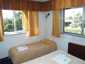 A single bed with a beige bedspread, two windows with yellow curtains, a wall-mounted light fixture, and a radiator. - Residencia Familiar La Rosa (San Carlos de Bariloche)