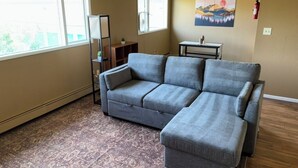 Ein Wohnzimmer mit einem grauen Ecksofa, einem gemusterten Teppich, einem kleinen Tisch und einem Bild an der Wand.