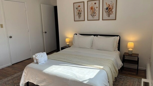Ein Schlafzimmer mit einem großen Bett, zwei Nachttischen mit Lampen, einer Tür und drei gerahmten floralen Kunstwerken an der Wand.