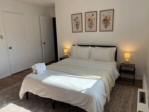 A bedroom with a large bed, two bedside tables with lamps, a door, and three framed floral artworks on the wall. - Newly Updated- Top Floor- Bona Nook #5 (Anchorage)