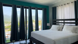 Een hotelkamer met een groot bed, een stadsgezicht door de vloerhoogte ramen en een plafondventilator.