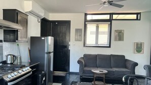 Een knus woongedeelte met een kitchenette, een grijze bank, een kleine ronde tafel en een plafondventilator.
