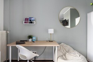 A bedroom with a bed, a desk with a chair, a round mirror, and a shelf with books.