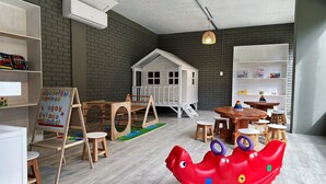 Una sala de jocs infantil amb una caseta, taules de fusta, cadires i un vaixell de joc vermell.