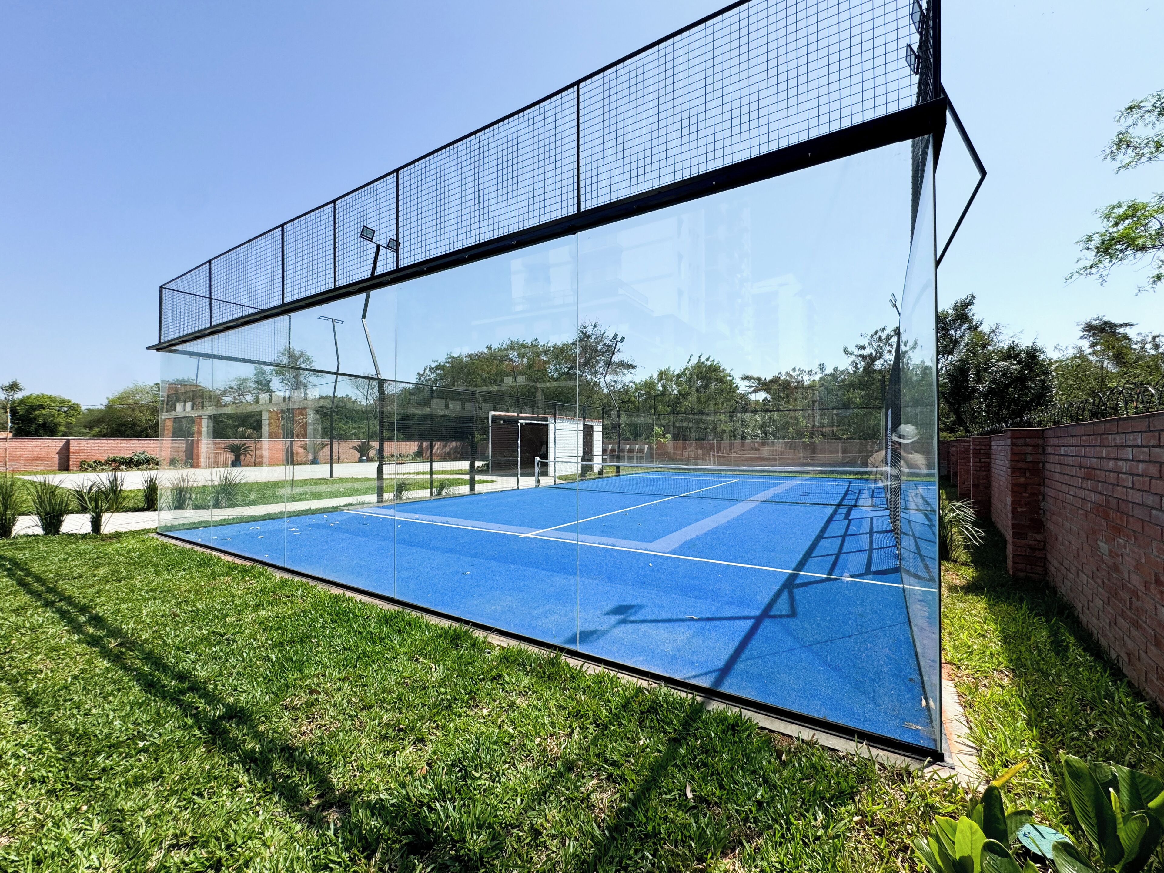 Cancha de tenis con cerramiento de vidrio, rodeada por un muro de ladrillo y vegetación.