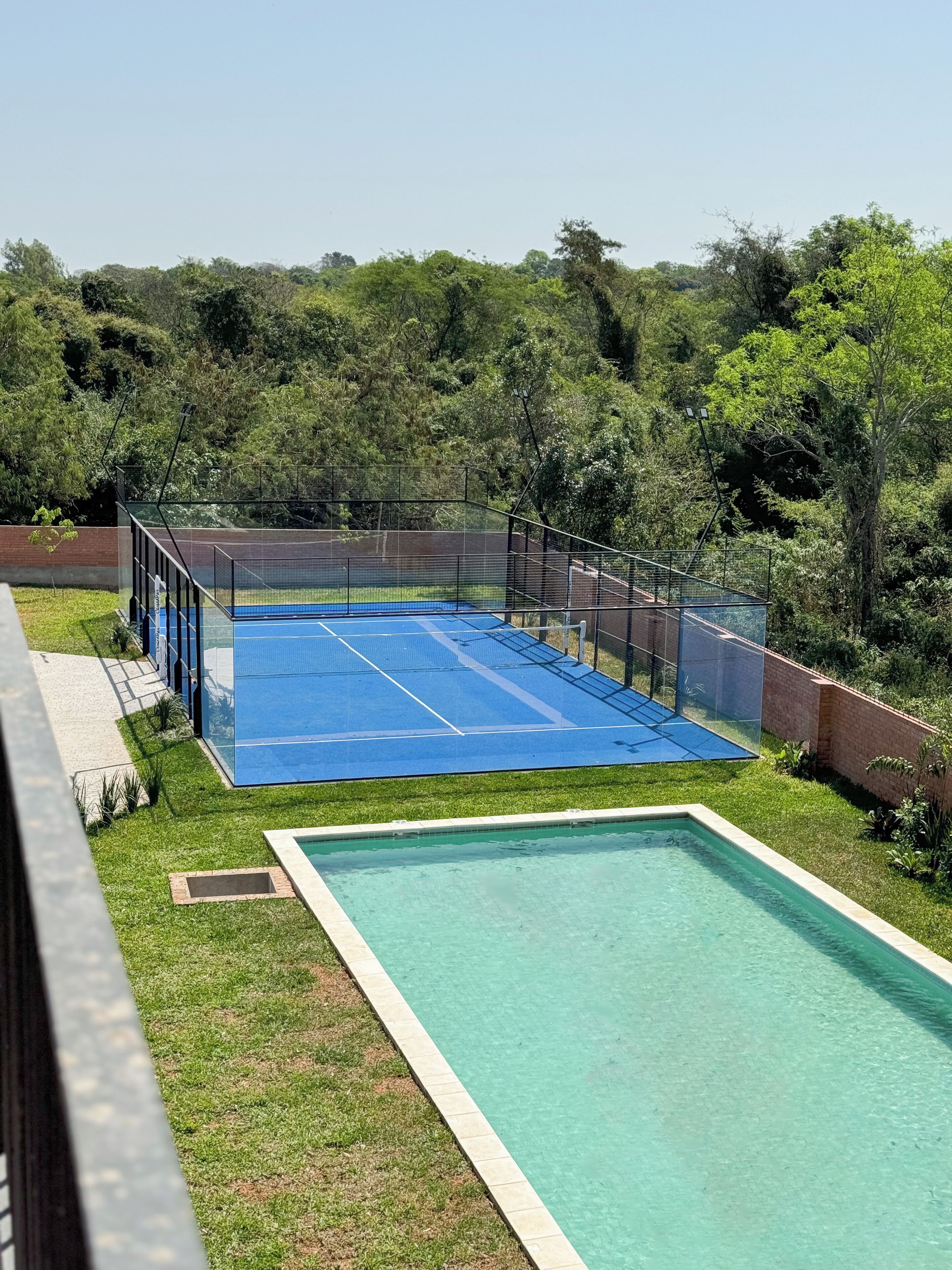 Una cancha de tenis con una cerca perimetral, una piscina y una zona verde con árboles de fondo.