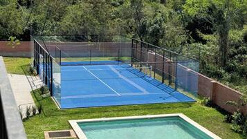 Una cancha de tenis con una cerca perimetral, una piscina y una zona verde con árboles de fondo.