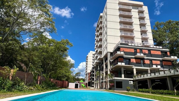 Una piscina amb vistes a un edifici d'alguns pisos i balcons, sota un cel blau clar.