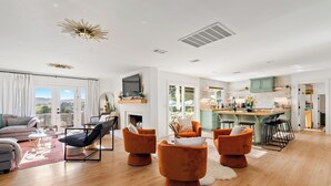 A spacious living room with a sofa, orange armchairs, and a coffee table. A kitchen with bar stools is visible in the background.