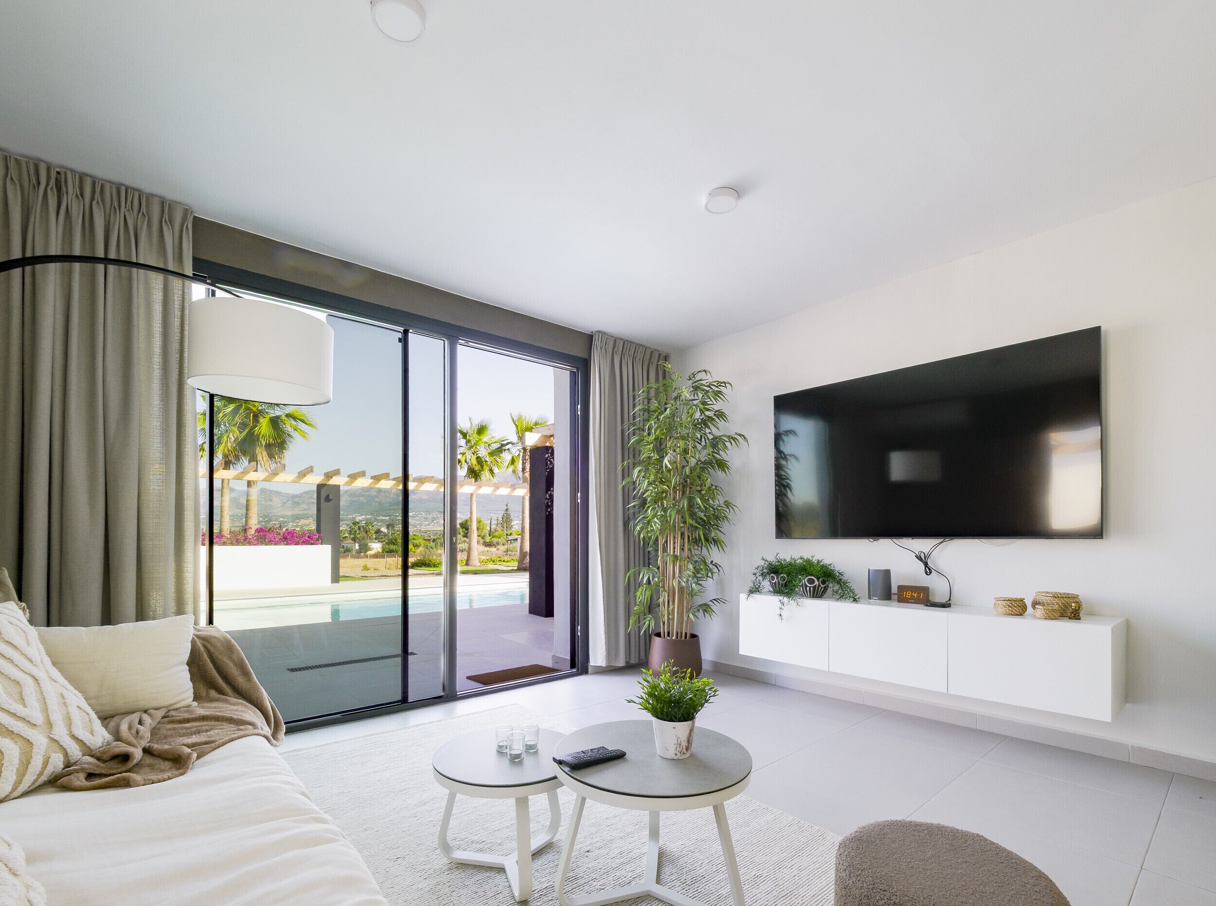 A modern living room with a large TV, a sofa, a coffee table, and a sliding glass door leading to a balcony with a pool view.