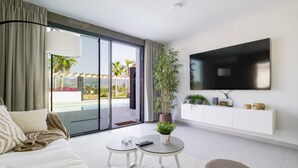 A modern living room with a large TV, a sofa, a coffee table, and a sliding glass door leading to a balcony with a pool view.