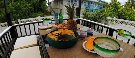 A table set with snacks, drinks, and a pineapple on a balcony with a view of a house and greenery.
