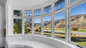 A bay window with a white cushioned bench, offering a view of a rural landscape with hills and a road. - Lakeside Retreat - Cromwell Holiday Home (Cromwell)