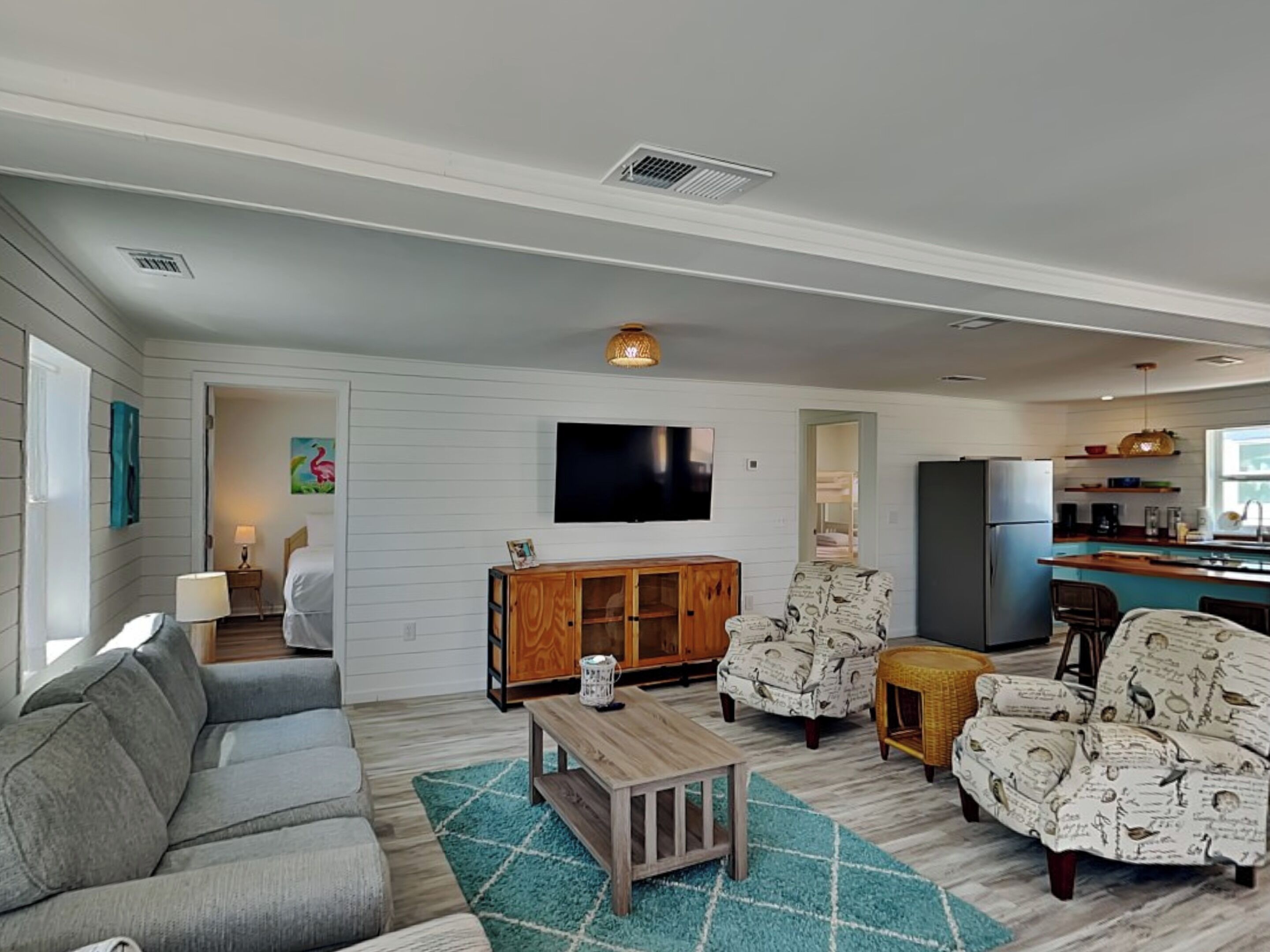 A living room with a gray sofa, a wooden coffee table, patterned armchairs, and a television on the wall.