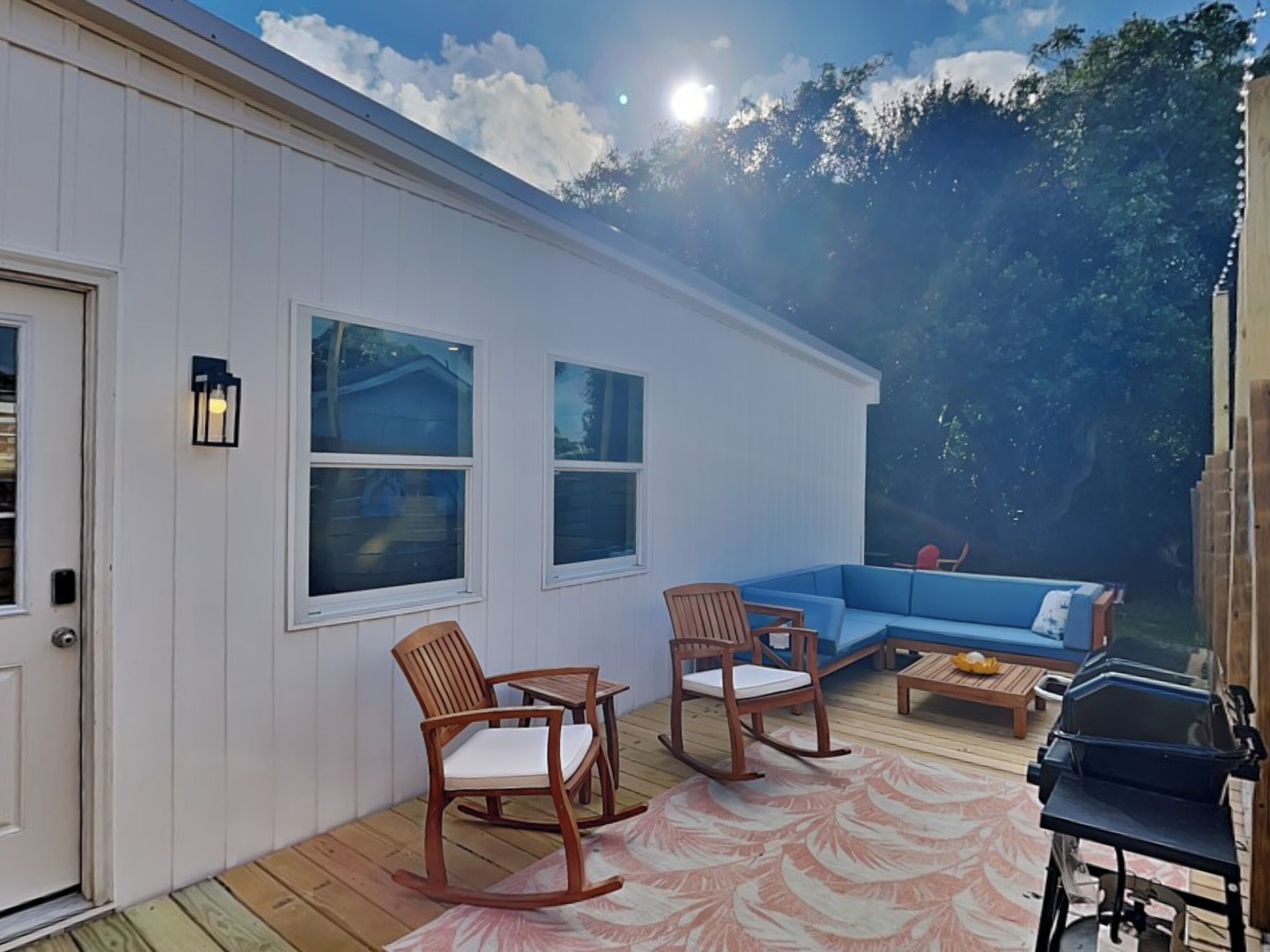 A covered patio with a blue sofa, two rocking chairs, a grill, and a wooden deck.