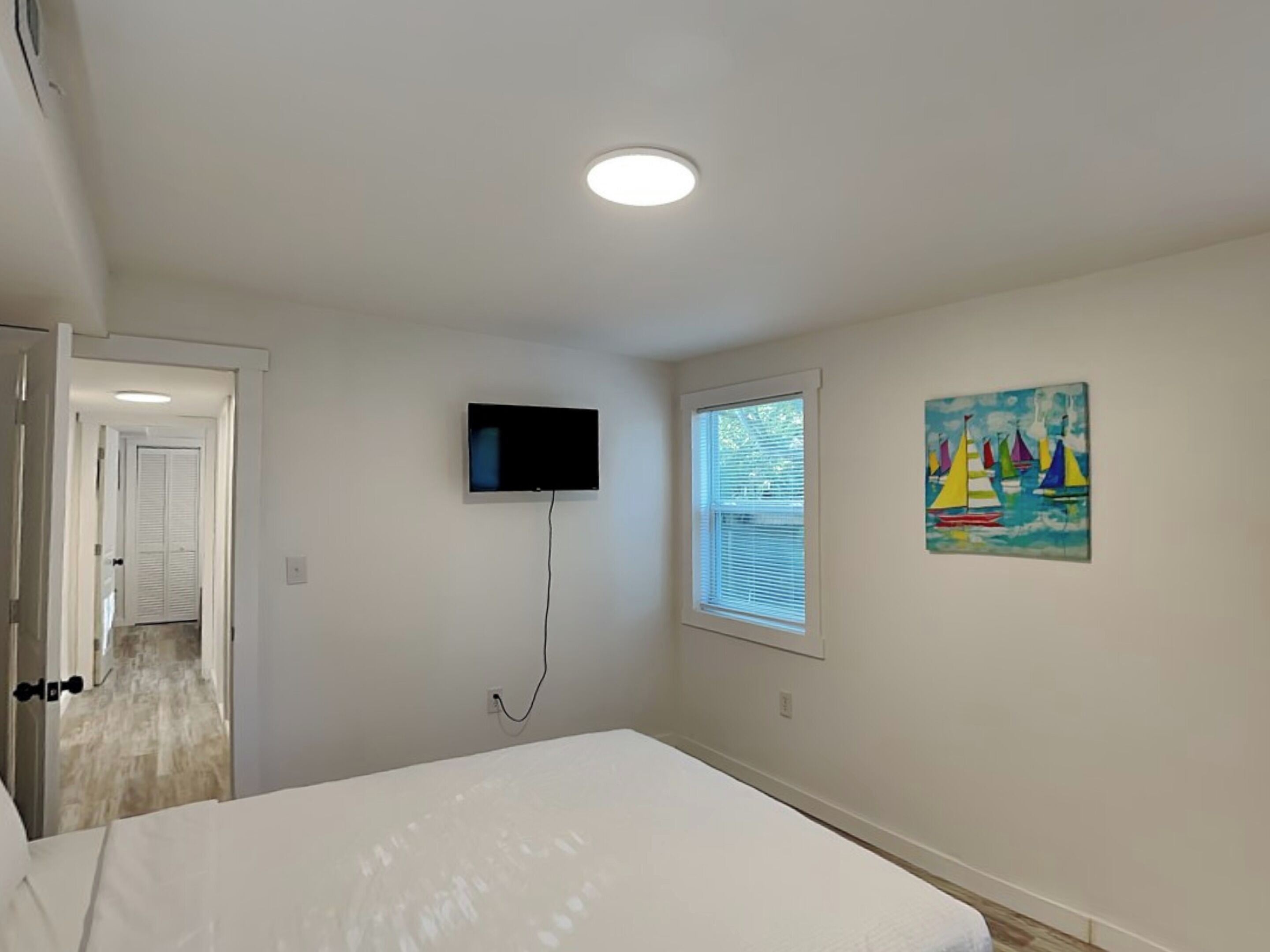 Une chambre à coucher avec un lit, un téléviseur fixé au mur, une fenêtre avec des stores et un tableau accroché au mur.