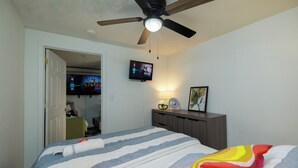 Een slaapkamer met een bed, een plafondventilator, een aan de muur bevestigde televisie, een nachtkastje met een lamp, en een deur naar een andere kamer.