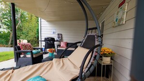 Eine überdachte Terrasse mit Bett, Stühlen, einem kleinen Tisch und einem Grill.