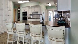 A kitchen with white cabinets, a central island with bar stools, stainless steel appliances, and recessed ceiling lighting. - Lion On The Beach by Resort Collection (Panama City Beach)
