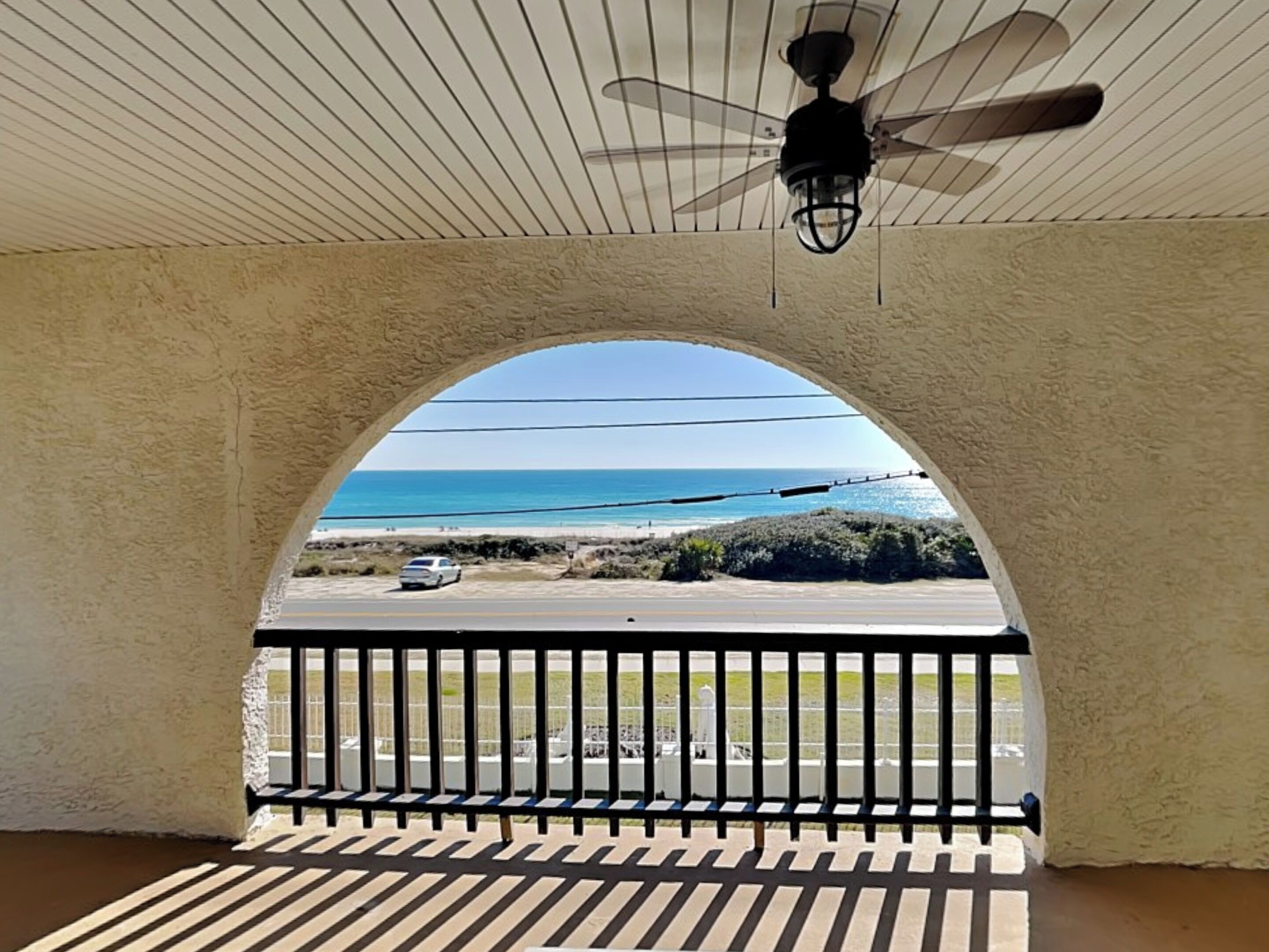 A covered outdoor area with a view of the beach, a ceiling fan, and a railing.