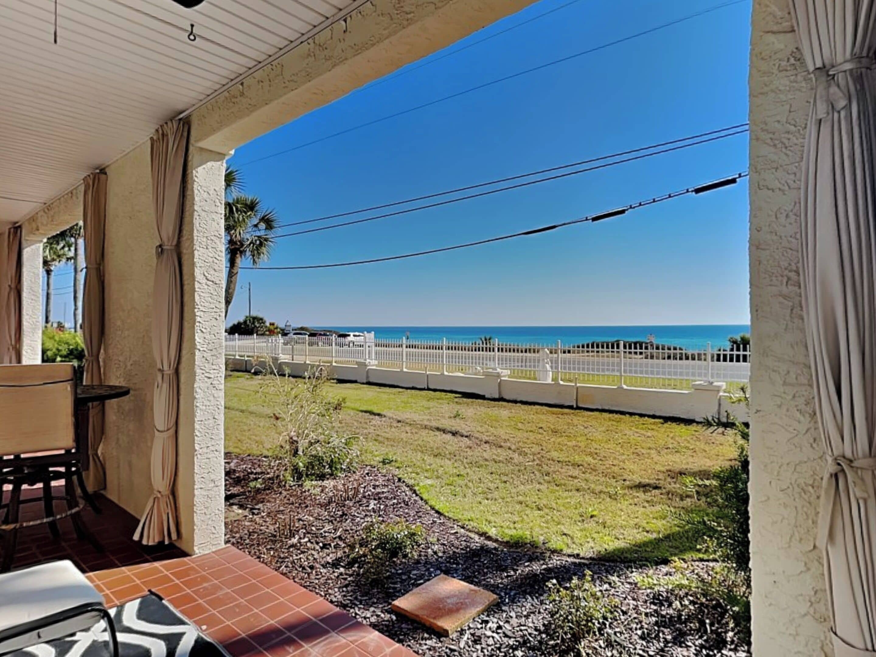 A covered patio with a view of the ocean, a white fence, and a grassy area.