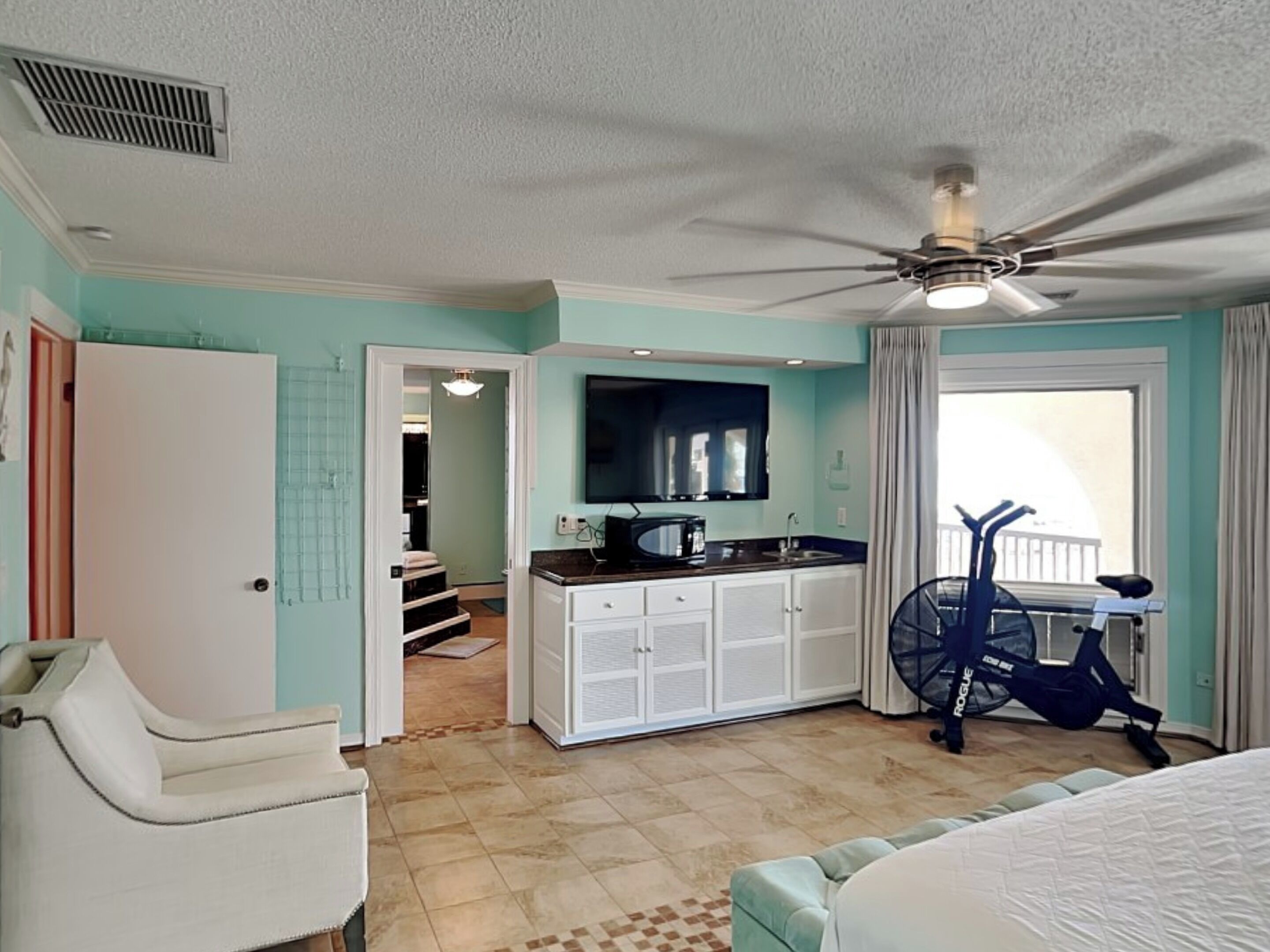 A compact hotel room with a kitchenette, a ceiling fan, a bed, and a window with curtains.