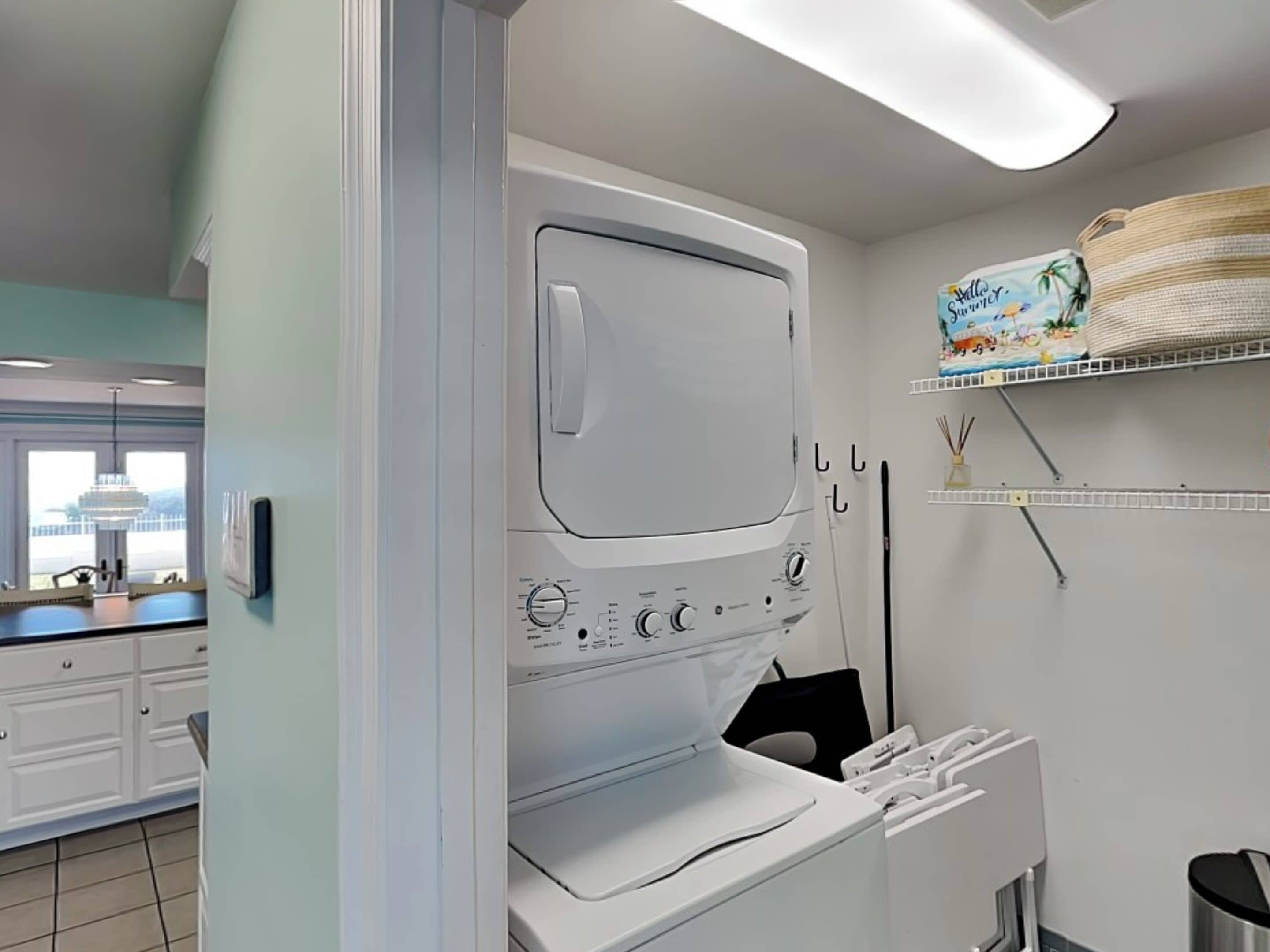 A modern laundry room with a large washer and dryer, a window with a view, and a shelf with a Hello Summer sign.