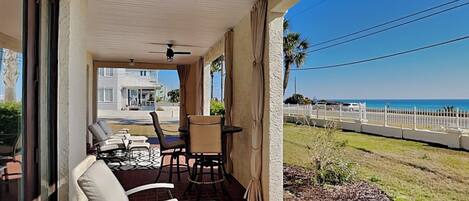 A covered patio with outdoor seating, a view of the ocean, and a white fence along the beachfront.