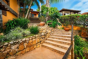 A stone staircase leading to a house with a tiled roof, surrounded by lush greenery and a clear sky. - Bz81 Pé na areia na Praia da Ferradura (Buzios)