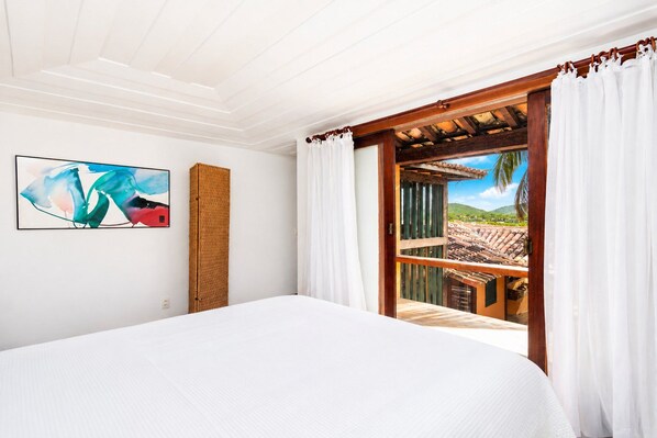 A hotel room with a bed, a painting, a wicker basket, and a view of a balcony with a wooden railing.
