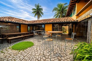 Un patio al aire libre con suelo de piedra, mesas y sillas de metal, bancos de madera y una pequeña zona de césped.