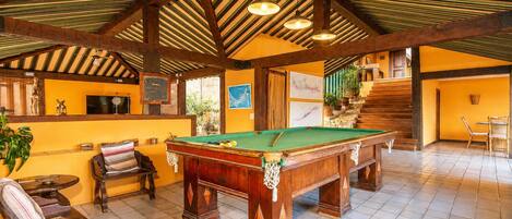 Una sala de billar con mesa de pool, muebles de madera y una escalera que sube a un nivel superior.