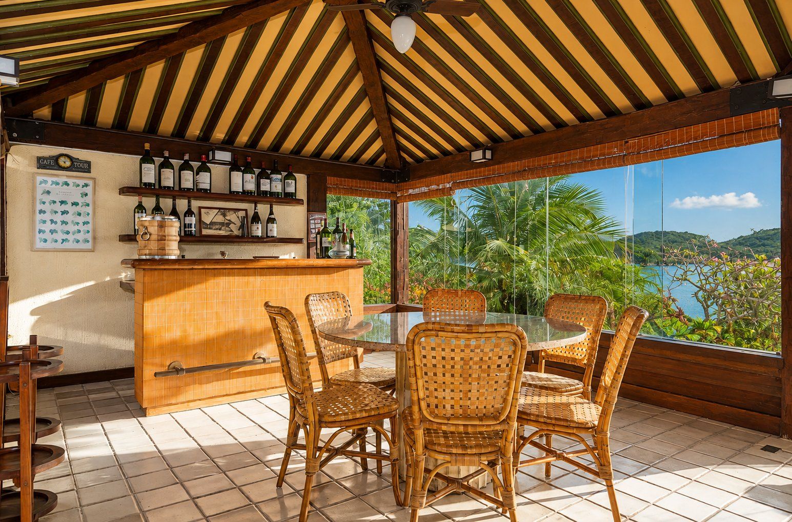 Un espacio de estar techado al aire libre con sillas de mimbre y una mesa de vidrio, con vistas a un paisaje con palmeras y colinas.