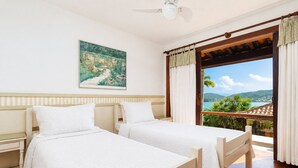 A hotel room with two beds, a ceiling fan, a painting, and a balcony with a view of the sea and hills. - Bz81 Pé na areia na Praia da Ferradura (Buzios)