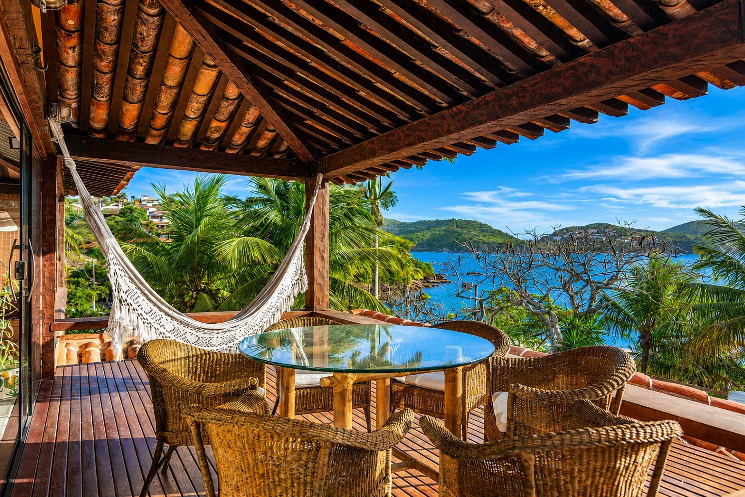 Un área de estar al aire libre con techo, amueblada con muebles de mimbre y una mesa de vidrio, con vistas a un paisaje costero con palmeras y el mar.