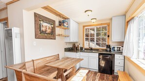 Een compacte keuken met een houten tafel en bankje, witte kastjes, en een raam met uitzicht op bomen.