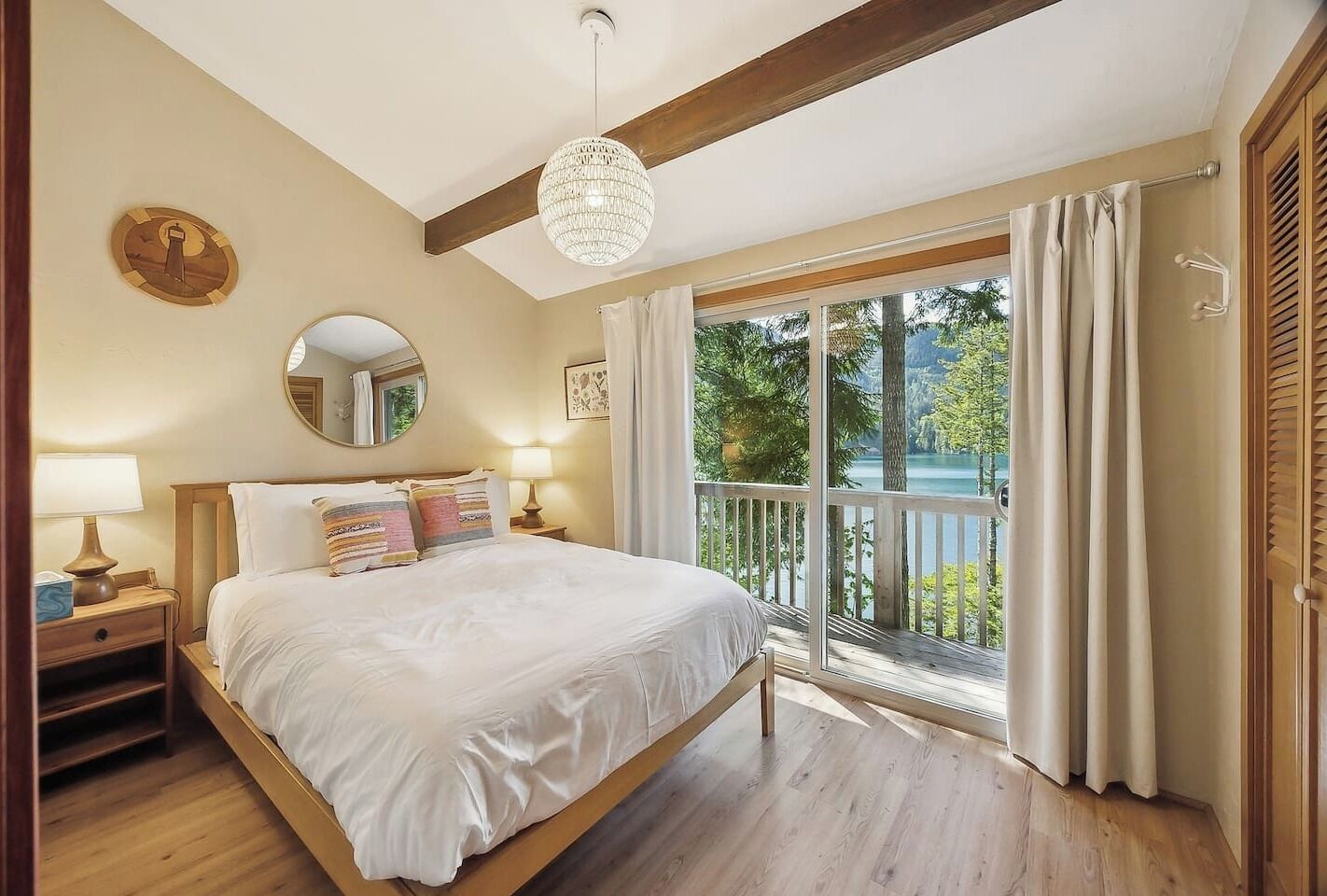A bedroom with a wooden bed, a round mirror, bedside lamps, and a balcony with a view of trees and a lake.