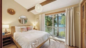 A bedroom with a wooden bed, a round mirror, bedside lamps, and a balcony with a view of trees and a lake.