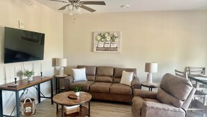 Ein Wohnzimmer mit braunem Ledersofa, einem runden Couchtisch, einem an der Wand montierten Fernseher und einem Deckenventilator.