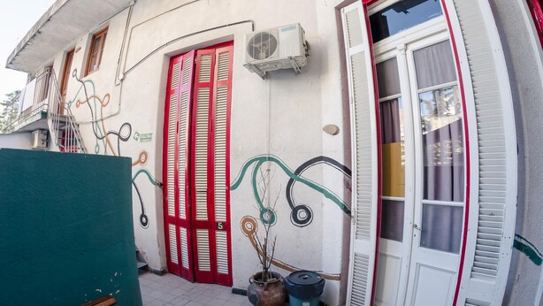 A building with a red door, a white door, and a window with shutters. There is a potted plant and a trash bin outside. - La Comunidad Hostel (Rosario)