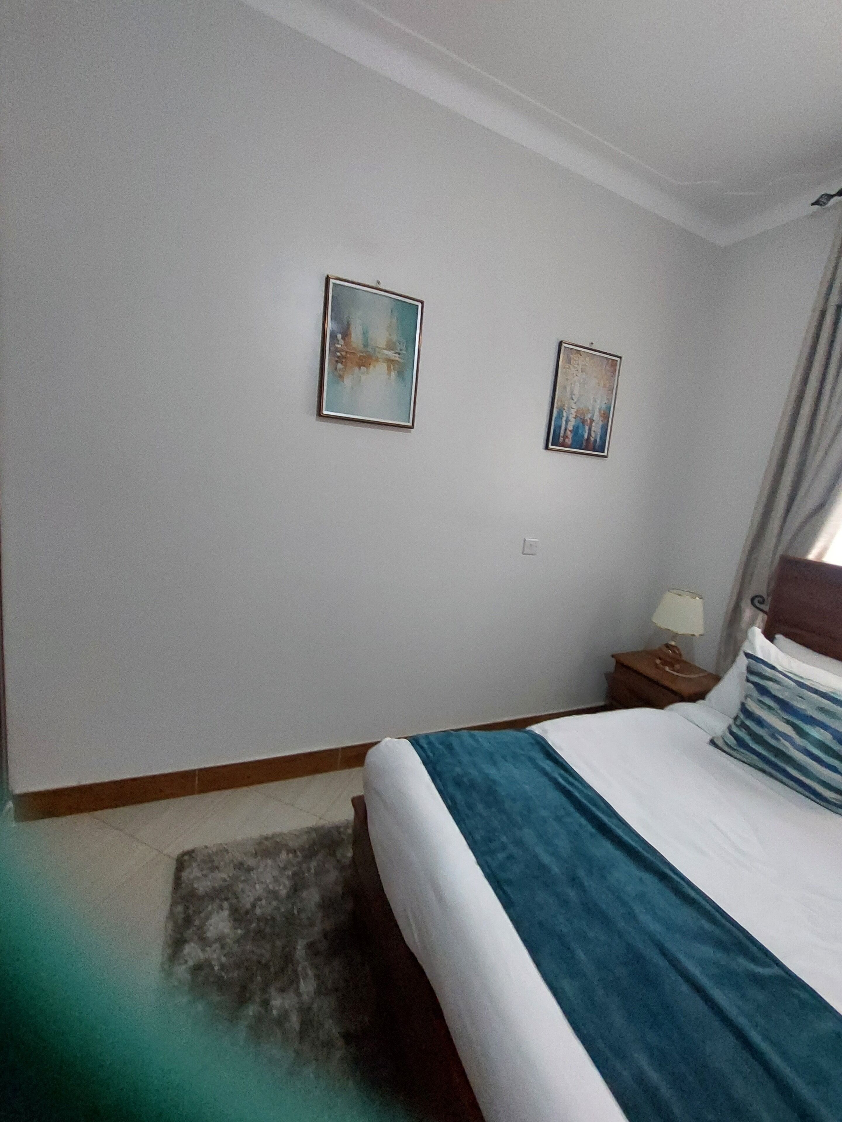 A hotel room with a bed, a nightstand, two wall-mounted paintings, and a window with curtains.