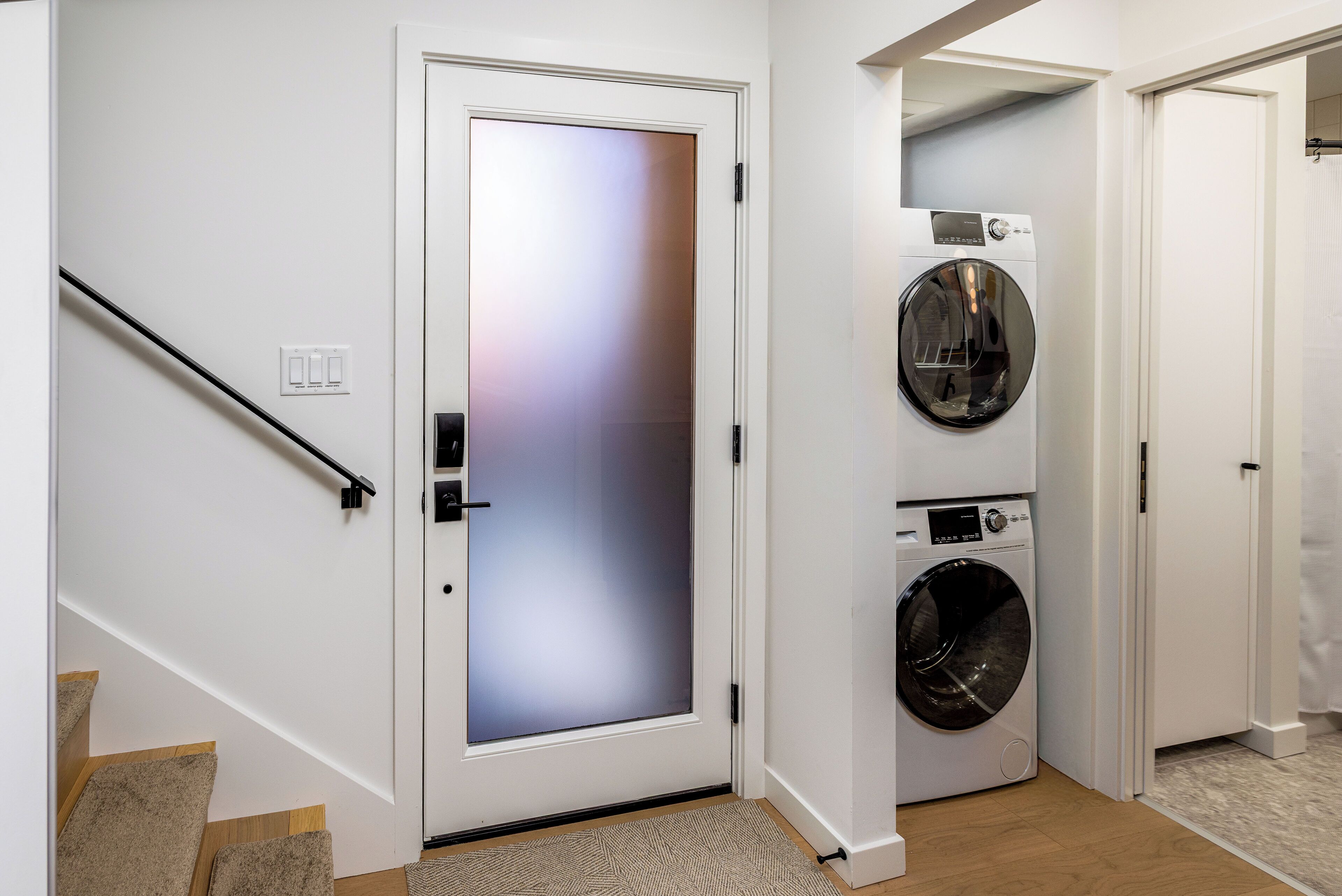 A laundry room with a front-loading washer and dryer, a door with frosted glass, and a staircase with carpeted steps.